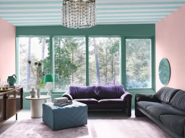 Ett vardagsrum med art deco inredning, mjuklila soffa, mörkgrå soffa, turkos pall och en stor fönsterutsikt över skogen.